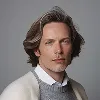 Portrait of a man with medium-length hair wearing a white sweater and gray collared shirt against a gray background.